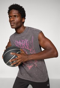 Ung mand i ærmeløs grafisk skjorte, der holder en sort og grå basketball, klar til at passe eller skyde mod en ensartet baggrund.