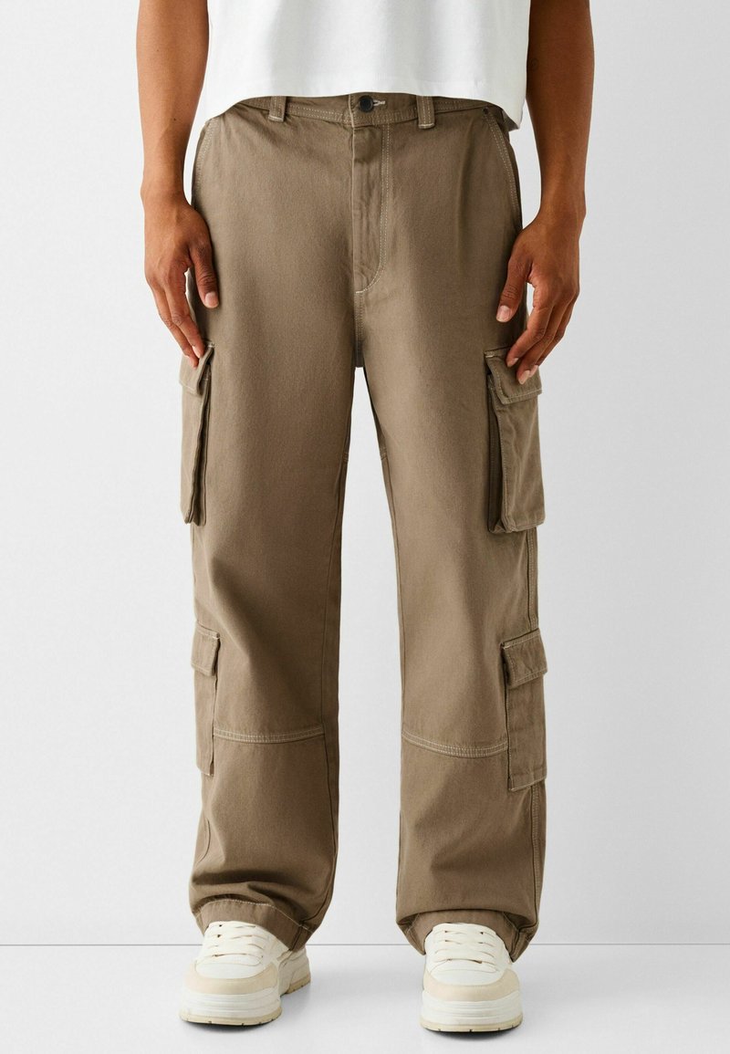 Pantalon cargo marron en coton, doté de multiples poches latérales, d'une coupe décontractée et de détails de couture blancs. Associé à des baskets blanches.