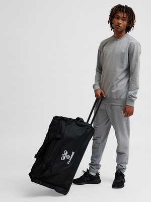 Młody mężczyzna w szarej dresie trzyma czarną walizkę na kółkach Hummel na białym tle.