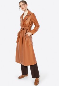 Brauner Leder-Trenchcoat mit tailliertem Gürtel, eingeschnalltem Kragen, langen Ärmeln und Seitentaschen. Kombiniert mit dunkelbraunen Hosen und Schuhen.
