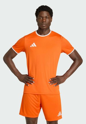 Mann steht mit den Händen in die Hüften gestützt, trägt ein leuchtend orangefarbenes Sportshirt und Shorts mit weißem Saum und Logo auf Brust und Bein.