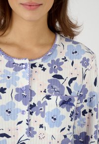 Blusa floral ligera en blanco, con patrones de flores azules y lavanda. Detalles destacados incluyen botones y un bolsillo con un acento de cinta.