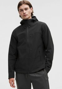 Svart softshell-jacka med huva, med fickor med dragkedja och en slät, matt yta. Den har en figursydd design och minimala sömmar.