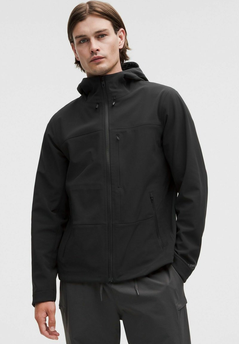 Svart softshell-jacka med huva, med fickor med dragkedja och en slät, matt yta. Den har en figursydd design och minimala sömmar.