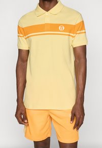 Geel poloshirt met oranje strepen, korte mouwen, twee-knops sluiting en een logo op de borst. Gecombineerd met bijpassende gele shorts.