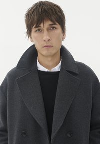 Ung man med kort brunt hår iförd en mörkgrå oversized kappa över en svart tröja och vit skjorta mot en enkel bakgrund.