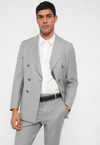 Giacca doppiopetto a righe grigie con pantaloni abbinati. Realizzata in tessuto liscio, presenta rever a lancia e bottoni in metallo. Camicia bianca sotto.