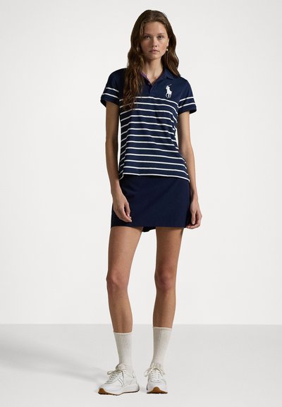 Marineblå stripete polo shirt med korte ermer, hvite horisontale striper, og en logo. Kombinert med et matchende marineblå skjørt og hvite joggesko.