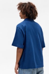 Camicetta a maniche corte con colletto in un blu profondo, tessuto testurizzato. Presenta una vestibilità rilassata e cuciture visibili lungo l'orlo.