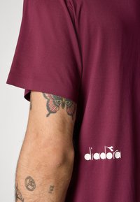T-shirt corto maroon in tessuto testurizzato, con il logo bianco "diadora" vicino all'orlo e un tatuaggio visibile sul braccio.