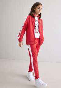 Red track jacket and pants with white side stripes, featuring a graphic t-shirt. Worn with white sneakers, set against a plain background.