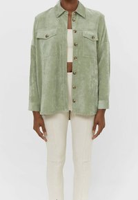 Green, textured jacket with a collar, front button closure, and two chest pockets. Model pairs it with a cropped top and light-colored pants.