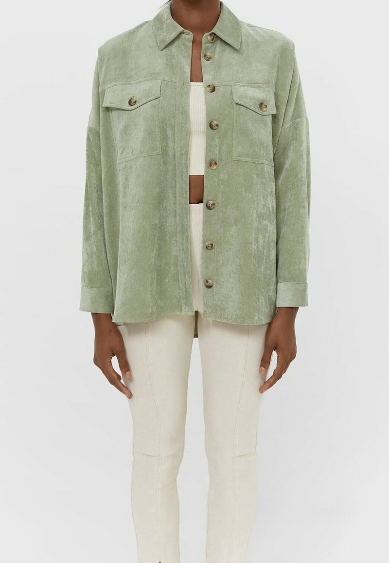 Green, textured jacket with a collar, front button closure, and two chest pockets. Model pairs it with a cropped top and light-colored pants.