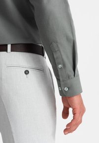 Chemise à manches longues grise avec une texture lisse, poignets fixés et boutons bleus, associée à un pantalon gris clair avec une poche arrière à bouton.