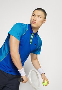 Joueur de tennis masculin en polo bleu se prépare à servir, tenant une raquette blanche et une balle jaune sur un fond uni.