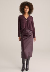 Plommonfärgad blus med textur, V-ringning och en dekorativ knapp, i kombination med en högmidjad plommonfärgad kjol i konstläder och knähöga stövlar.