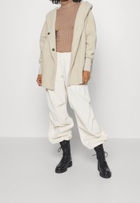 Beige fleecejacka med huva och två svarta knappar, ribbad brun polotröja, vita löst sittande byxor, svarta snörade ankelstövlar.