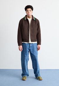 Brun uldblandingsjakke med knaplukning, grøn og hvid stribet krave, parret med lyseblå baggy jeans og sneakers med grønne kanter.