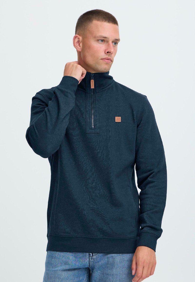 Moški s kratkimi lasmi, oblečen v temno modro pulover z četrt zadrgo in svetlo modre kavbojke, ki si prilagaja ovratnik in gleda v desno na enobarvni beli podlagi.