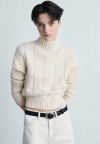 Cremefarbenes Strickpullover mit hohem Kragen und gerippten Bündchen, kombiniert mit einem schwarzen Gürtel über einem hellen Rock. Strukturierte Garn-Details.