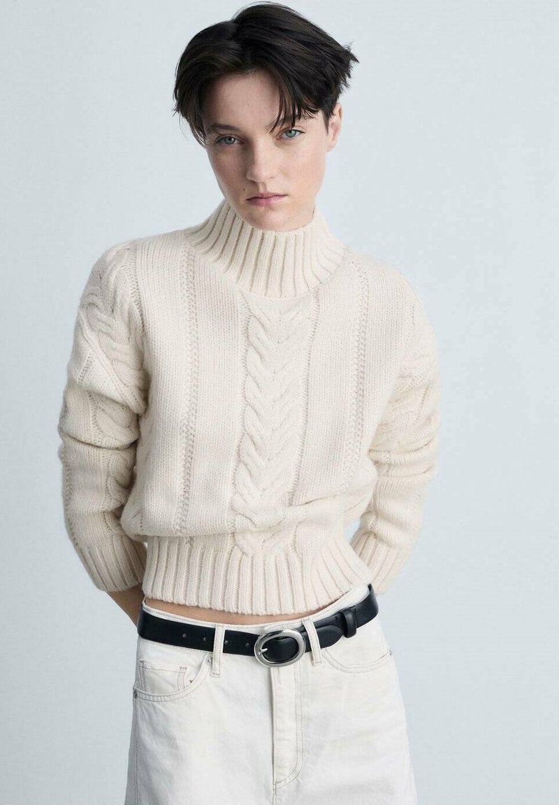 Cremefarbenes Strickpullover mit hohem Kragen und gerippten Bündchen, kombiniert mit einem schwarzen Gürtel über einem hellen Rock. Strukturierte Garn-Details.