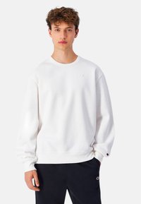 Sudadera blanca de cuello redondo en tejido suave, con un ribete en el cuello y en los puños, y un pequeño detalle de logo en el pecho. Combinada con pantalones oscuros.