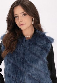 Blå faux fur-väst med hög krage, med ett mjukt, texturerat tyg och en dold framknäppning. Buren över en svart topp.
