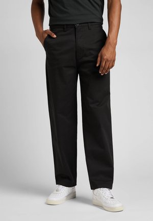 Person trägt schwarze Hose mit geradem Bein, schwarzes Hemd, weiße Sneaker, mit einer Hand in der Tasche vor einfachem Hintergrund.