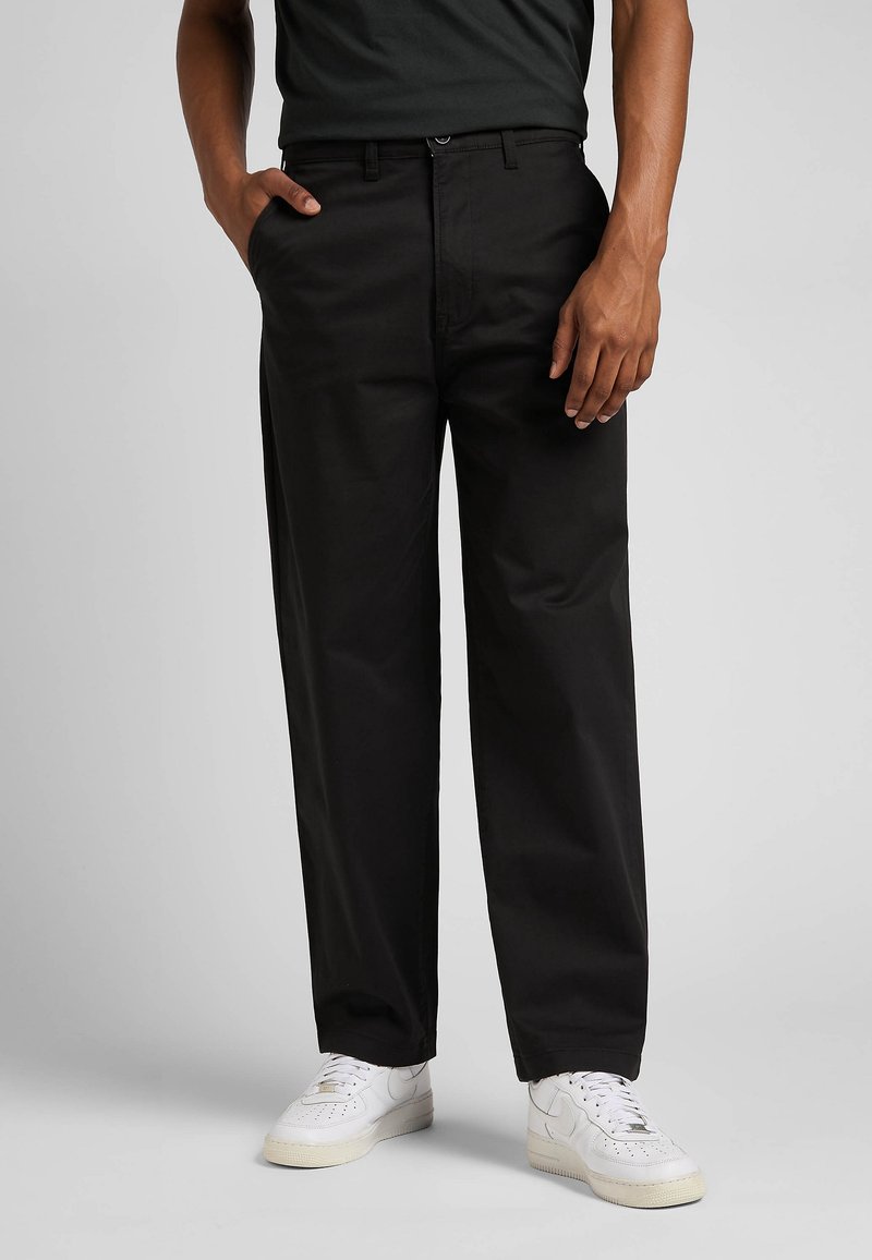 Personne portant un pantalon noir droit, une chemise noire, des baskets blanches, une main dans la poche devant un fond uni.