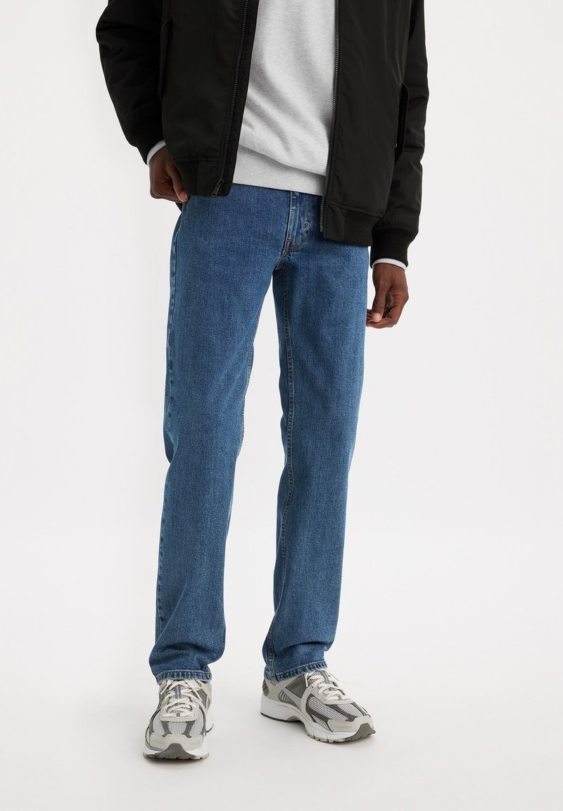 Jeans de mezclilla azul con corte recto, que presentan cinco bolsillos, costuras sutiles y una textura desgastada, combinados con una chaqueta bomber negra y zapatillas.