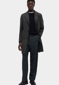 Cappotto di lana grigio con colletto a punta, maglione nero e pantaloni a righe. Texture liscia e vestibilità su misura, con toni neutri.