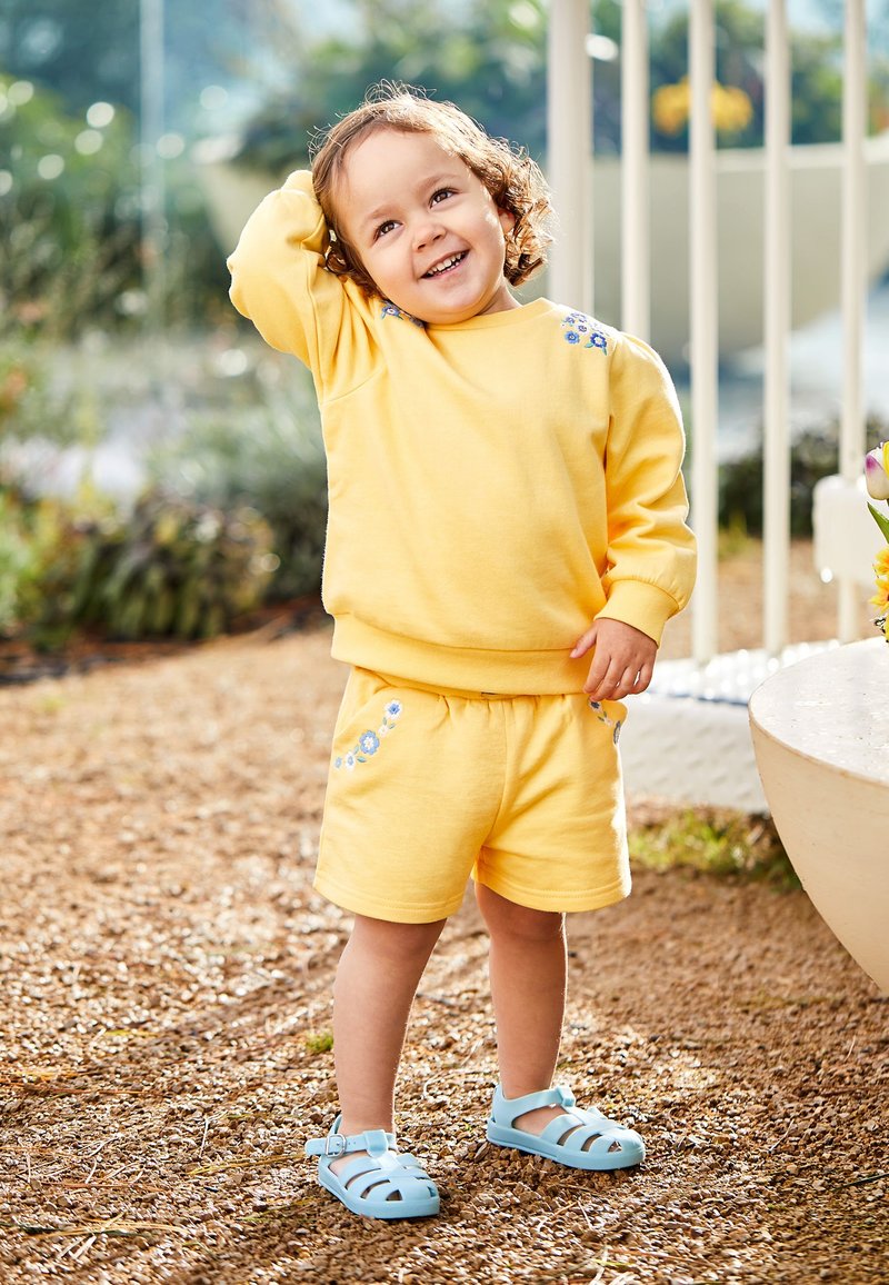Niño pequeño con sudadera amarilla y pantalones cortos con flores azules, de pie al aire libre sobre grava, sonriendo y tocándose la cabeza.