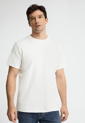 Uomo che indossa una maglietta bianca a maniche corte tinta unita e jeans blu, in piedi davanti a uno sfondo grigio chiaro.