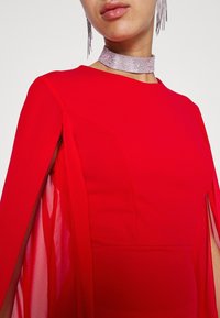 Femme portant une robe rouge vif avec des manches cape transparentes et un collier ras-du-cou argenté étincelant avec des boucles d'oreilles pendantes assorties.
