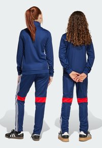 Los chándales deportivos de la marina presentan mangas largas, un cuello alto y acentos a rayas; los pantalones tienen bandas rojas audaces. Tejido texturizado y ligero.
