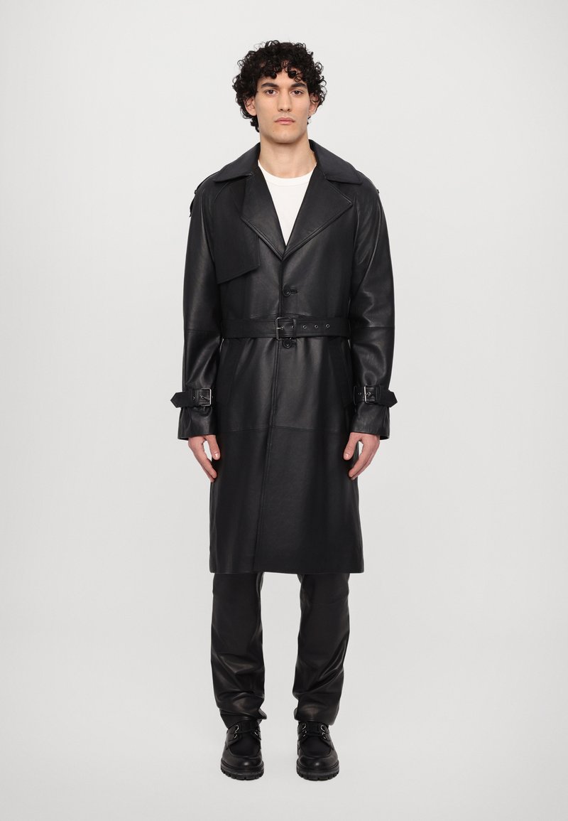Man med lockigt hår iförd lång svart läder trench coat, svarta läderbyxor, svarta stövlar och vit skjorta, står mot en neutral bakgrund.