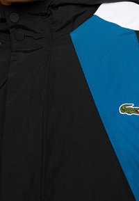Czarna i turkusowa wiatrówka z teksturowanej tkaniny, z zapięciem na zatrzaski i logo krokodyla Lacoste na dolnej prawej stronie.
