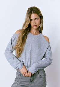 Top gris de manga larga con hombros al aire, con acentos de botones plateados en los hombros. Llevado con jeans gris de cintura alta.