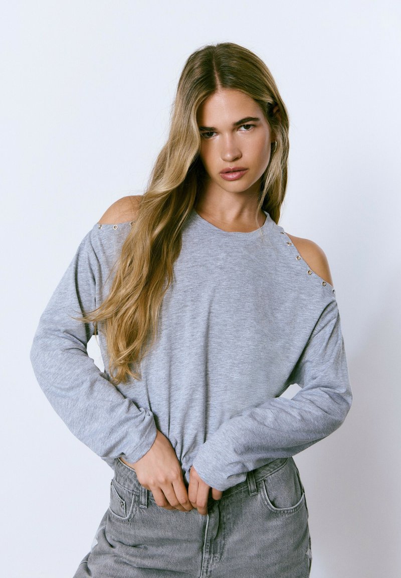 Top gris de manga larga con hombros al aire, con acentos de botones plateados en los hombros. Llevado con jeans gris de cintura alta.