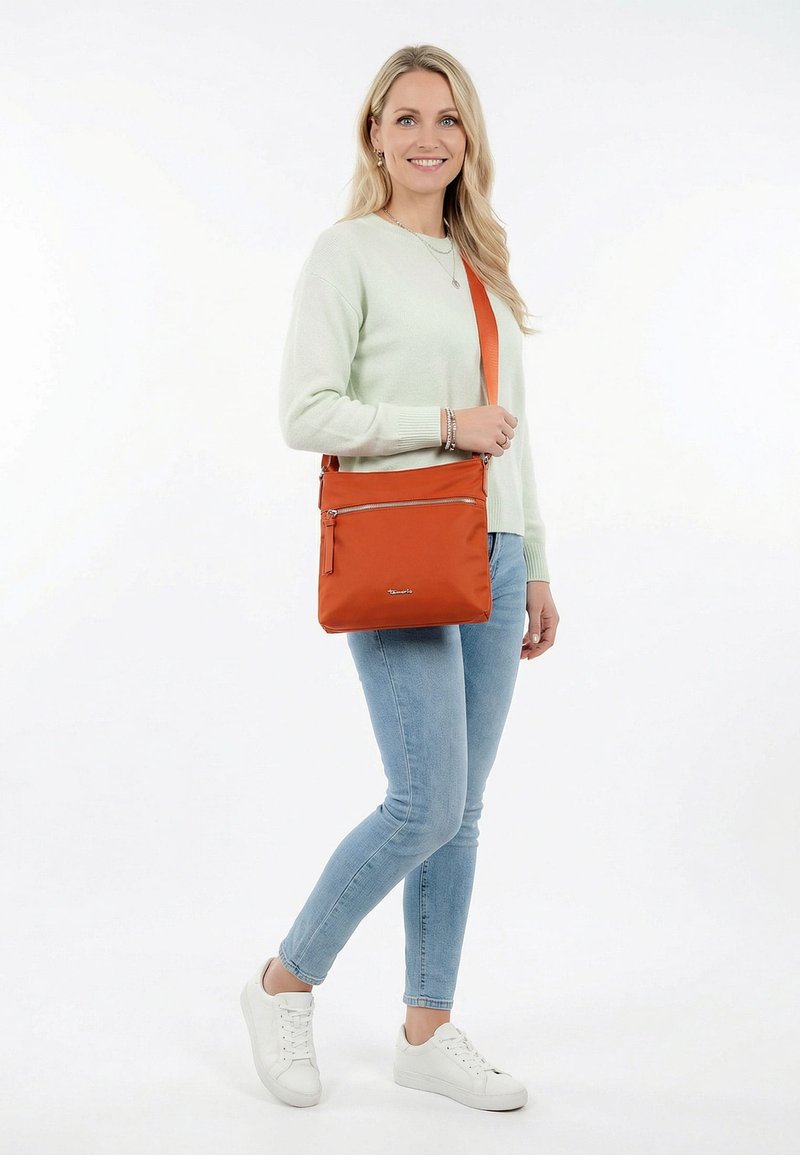 Frau steht lächelnd da, trägt einen leichten Pullover, Jeans und weiße Turnschuhe, hält eine orangefarbene Umhängetasche mit vorderer Reißverschlusstasche vor weißem Hintergrund.