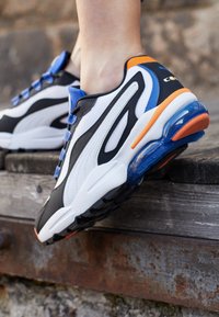 Baskets de sport noires, blanches et bleues en mesh et caoutchouc, dotées d'un coussin d'air visible, d'accents orange et de détails texturés.