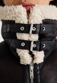 Braune Lederjacke mit plüschigem hellbeigen Lammfellkragen, ausgestattet mit silbernem Zubehör und zwei schwarzen Lederbändern mit Metall-Schnallen.
