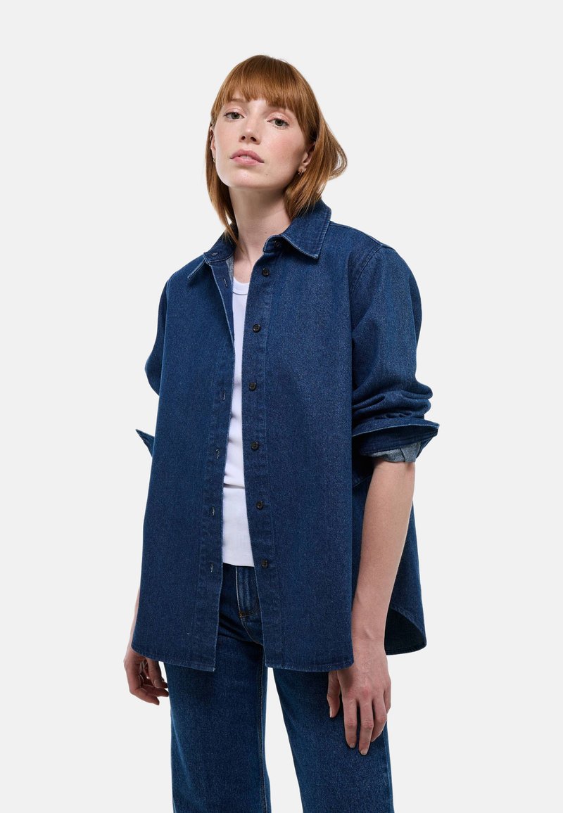 Chemise en denim bleu foncé, avec col pointu, manches longues, fermeture par boutons, et coupe ample. Associée à un débardeur blanc.