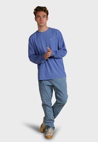 Mladý muž stojící s překříženýma nohama, oblečený v modré košili s dlouhým rukávem, světle modrých džínách a béžových teniskách, na čistém bílém pozadí.