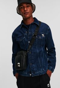 Jachetă din denim de culoare albastru închis cu �închidere cu nasturi și două buzunare pe piept, asociată cu o geantă umăr neagră ce are un motiv grafic.