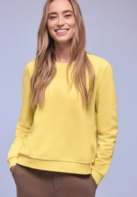 Jeune femme aux longs cheveux blonds portant un sweat-shirt jaune vif et un pantalon marron, souriant avec les mains dans les poches devant un fond clair.