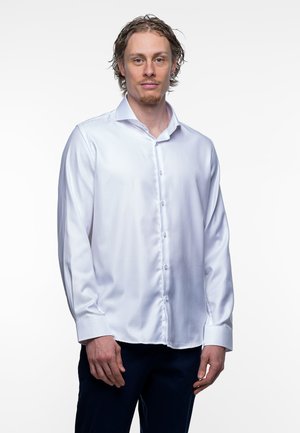 Man met licht krullend haar en een verzorgde baard, draagt een wit overhemd met knopen en donkere broek, staand voor een effen witte achtergrond.