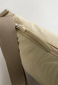 Beige Stofftasche mit einer strukturierten Oberfläche, ausgestattet mit einem oberen Reißverschluss und einem breiten braunen Tragegurt.