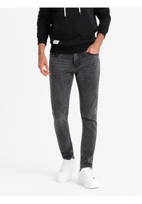 Jeans de mezclilla gris ajustados con desgastado sutil, cinco bolsillos y costuras en contraste, combinados con zapatillas blancas y una sudadera negra con capucha.