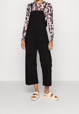 Person iført sort bredbenet overall, en langærmet abstrakt mønstret top i beige og sort samt cremefarvede sandaler med åben tå.